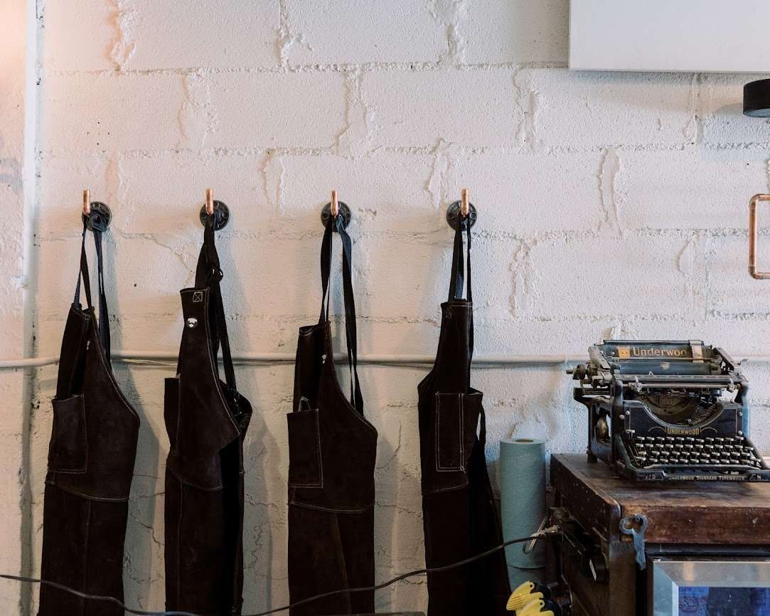 Black aprons hung up on metal hooks against white brick wall with black vintage typewriter in the corner