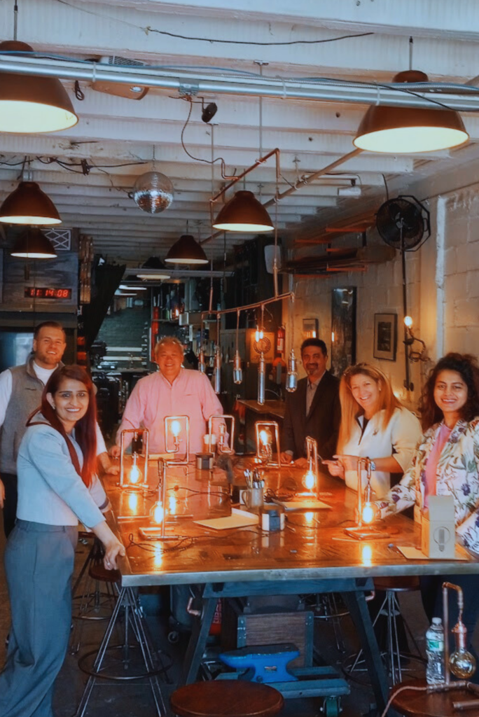 A group of people celebrating a birthday party in Brooklyn stand around a table smiling with their new crafts