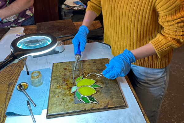 A person in a yellow shirt wearing blue gloves works on creating homemade stained glass creations for holiday gifts